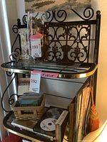 Side view of black metal bakers rack showing decorative back panel, glass shelves with gold trim, and various contents placed on shelves including glass vase, baskets, and CDs (contents not included).
