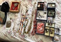 Full spread showing costume jewelry laid out on white and pink floral fabric. Includes necklaces, watches, clip-on earrings, bracelets, and brooches arranged in boxes and loose on fabric.