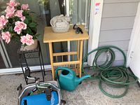Shows faux pink roses in a pot on metal stand, wooden table with stone planter, green watering can, green garden hose, blue propane tank on cart, and hose attachments.