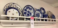 Photo showing collection of blue and white decorative plates arranged on a white wall-mounted wooden shelf with guard rail and turned spindle supports.
