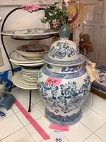 Large Asian style ceramic urn with lid and floral designs on tiled surface next to a metal plate holder stand with plates.