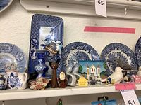 Blue and white decorative plates of various shapes and sizes arranged on a white shelf with various small figurines and pitchers.