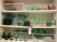 Wide view of three shelves with green, pink, and clear glassware including plates, cups, stemware, and bowls, plus some tableware and collectibles.