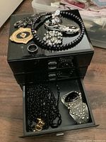 Black glossy acrylic jewelry box with assorted jewelry on top and inside the open drawer, showing various pieces such as a black bead necklace, rings, brooches, and pendants.