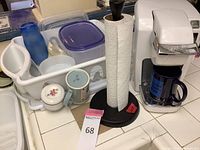White Keurig coffee maker with coffee mug, food containers, paper towel holder and other kitchen items arranged on counter.