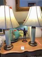 Photo of two Royal Haeger lamps with green ceramic and brass bases and cream colored shades on a wooden surface alongside sculptures and paintings