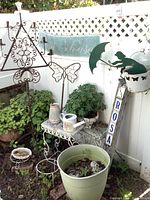 Photo shows a garden corner with black metal candle holder, white metal table, potted green plants, watering can and decorative garden sign