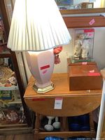 Photo showing the wooden drop leaf table with a broken leaf holder and the pink ceramic lamp with white pleated shade on top.