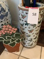 Full view of ceramic umbrella stand decorated with multicolor butterflies, showing multiple items inside and a decorative flower shaped clay or ceramic flower pot next to it.