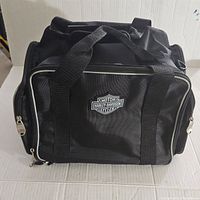 Front angled view showing Harley Davidson logo on front pocket with double carry handles and zippered compartments.