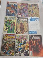 Photo showing nine vintage Avengers comic books laid in three rows on cardboard background, featuring various cover art with characters and team battles.