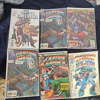 Photo showing 6 Captain America comics laid out on a surface, featuring diverse comic covers including a woman sitting and Captain America fighting villains.