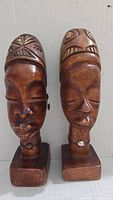 Front view of two vintage hand carved wooden heads mounted on square bases, showing detailed facial features and headdress patterns.