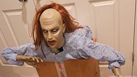 Prop figure of a possessed woman with red hair, pale face, and light blue nightgown, shown standing in a cardboard box.