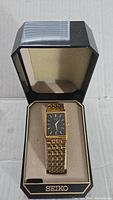 Gold-tone rectangular Seiko wristwatch with black dial and gold markers inside beige cushioned Seiko branded watch box.