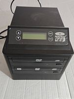 Front view of Zipspin DVD duplicator showing LCD screen, control buttons, and two DVD drives labeled DVD-ROM and DVD-RW.