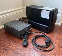 Photo showing three APC UPS battery units arranged on hardwood floor, visible black rectangular bodies with cables.