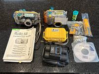 Lot of two Canon digital cameras in transparent waterproof housings with visible blue and yellow locking parts, located on countertop along with manuals, Pelican 1010 micro case, black Bushnell binoculars, and accessories.