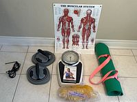 Photo showing all fitness items arranged on the floor: muscular system chart, analog scale, black push-up bars, green yoga mat with pink strap, yellow resistance band, skipping rope, and black cord.