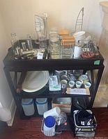 Wide view of kitchen lot items on a black shelf including plates, glass containers, cups, and various kitchen utensils.