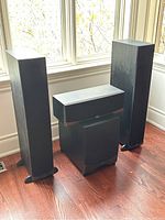 Overall lot showing two tall RF-15 tower speakers, the RC-25 center channel speaker in front resting on the black RW-10 subwoofer, positioned in front of windows on a wood floor.