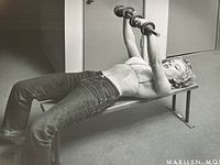 Close-up side view of Marilyn Monroe bench pressing dumbbells in black and white.