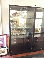 Front view of Michael Weiss black glass front display cabinet showing glass shelves with glassware inside and cupboard drawers below.