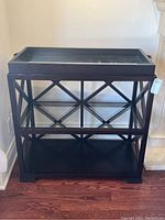Front view of black modern console table with a removable serving tray on top, showing three shelves and geometric X design on sides and back. Serving tray has dark handles with silver-tone accents.