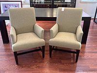 Pair of beige upholstered armchairs with dark wood legs shown in front of a dining table on a wooden floor.