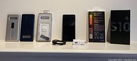Overview of all lot contents on shelf: three phone cases (silver sliding kickstand, navy ribbed, clear sealed), Samsung Galaxy S10+ smartphone, USB cable, Samsung fast charger, tempered glass screen protector box, S10+ retail box