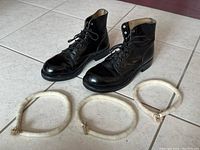 Pair of black leather high-top parade boots with metal foot pads, size 5.5, displayed on tiled floor with three white fabric parade pant weights in front.