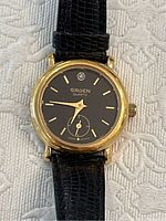 Front view of Gruen ladies quartz wristwatch showing black dial, gold-tone case, and textured black leather strap with buckle.