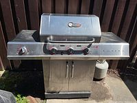 Front view of stainless steel Char-Broil BBQ grill with three knob controls, built-in thermometer on hood, and propane tank attached on right side.
