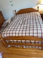 Full view of wooden bed frame made of light brown wood with plaid bedding showing headboard and footboard. Metal rails visible under footboard.