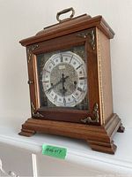 Front angled view of wooden Hamilton mantle clock showing brass handle on top, decorative accents, and clock face with Roman numerals.