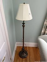Full view of floor lamp next to a sofa and wall, showing cream fabric shade and dark metal base and pole.