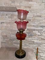 Front view of the entire glass oil lamp showing metal base, red glass reservoir, and two pink glass shades stacked above.
