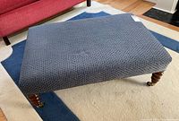 Rectangular dark gray fabric upholstered ottoman on light carpet with wooden turned legs and brass casters, photographed from above showing the top and one side.