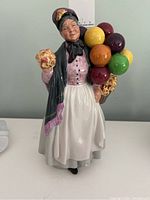 Front and side view of Royal Doulton Biddy Pennyfarthing HN1843 figurine showing woman in traditional dress holding a bouquet and a basket with colorful balloons.