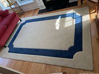 Full view of rectangular rug on hardwood floor near a red chair and furniture. Shows two-tone design with blue border and cream center.