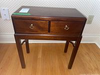 Front view of a wooden side table showing two drawers with brass metal handles and four supporting legs on a hardwood floor.
