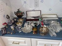 Full lot of silver-plated flatware, serving pieces, salt and pepper set, decorative spoons, and miscellaneous items spread on counter top.