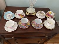 Eight teacups with matching saucers displayed on wood dresser surface, diverse floral patterns and colors visible.
