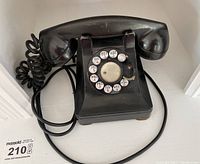 Front view showing black rotary dial telephone with handset on cradle, coiled cord, and visible signs of wear.
