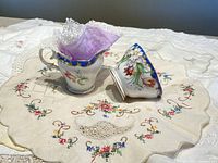 Bell China cream jug and sugar bowl with purple lace doily inside cream jug, placed on round cream-colored embroidered doily with floral motifs