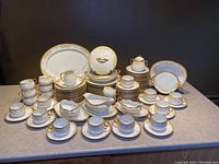 Full set arranged on countertop showing plates, saucers, cups, serving dishes with gold trimmed floral design