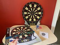 Electronic dart board displayed upright with digital display at bottom center, two sets of darts in plastic bags and instruction manual on table, original box upright beside board.
