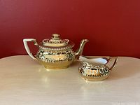 Teapot and creamer set displayed on light wood surface with red background showing detailed gold and cream patterns.