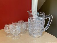 Full set of clear hobnail glass including one large pitcher and six stemmed juice cups arranged on table against red background.