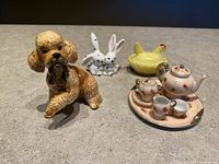 Photo of all items together showing the dog figurine, rabbits, yellow chicken and the tea set on a gray surface